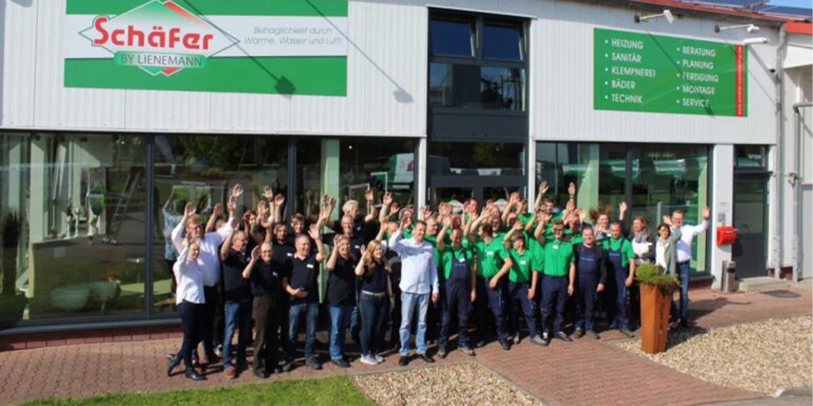 Eine große Gruppe von Mitarbeitern steht vor dem Firmengebäude und winkt fröhlich in die Kamera, um ihre Teamarbeit zu feiern.